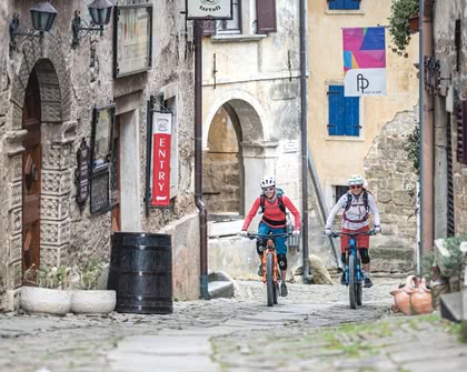 Two mountain bikers biking along an alleyway past stone buildings